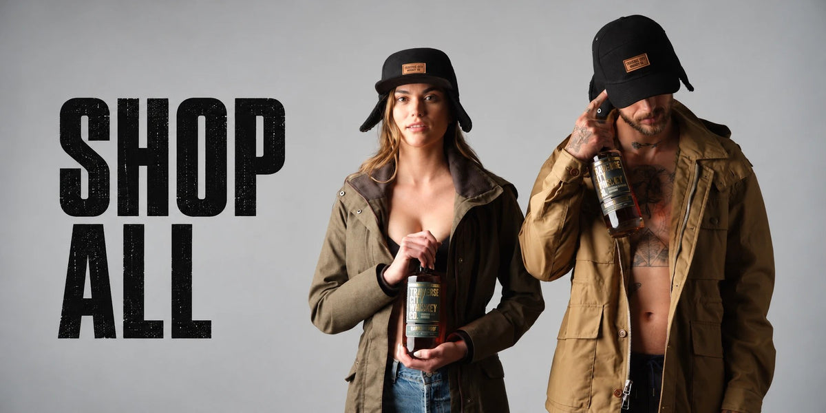Two models in Traverse City Whiskey Co. caps and canvas jackets holding bottles of whiskey, next to the words Shop All