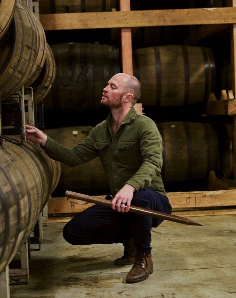A man in an olive green shirt and dark pants crouches in a wooden barrel room, holding a long wooden tool next to large oak barrels.