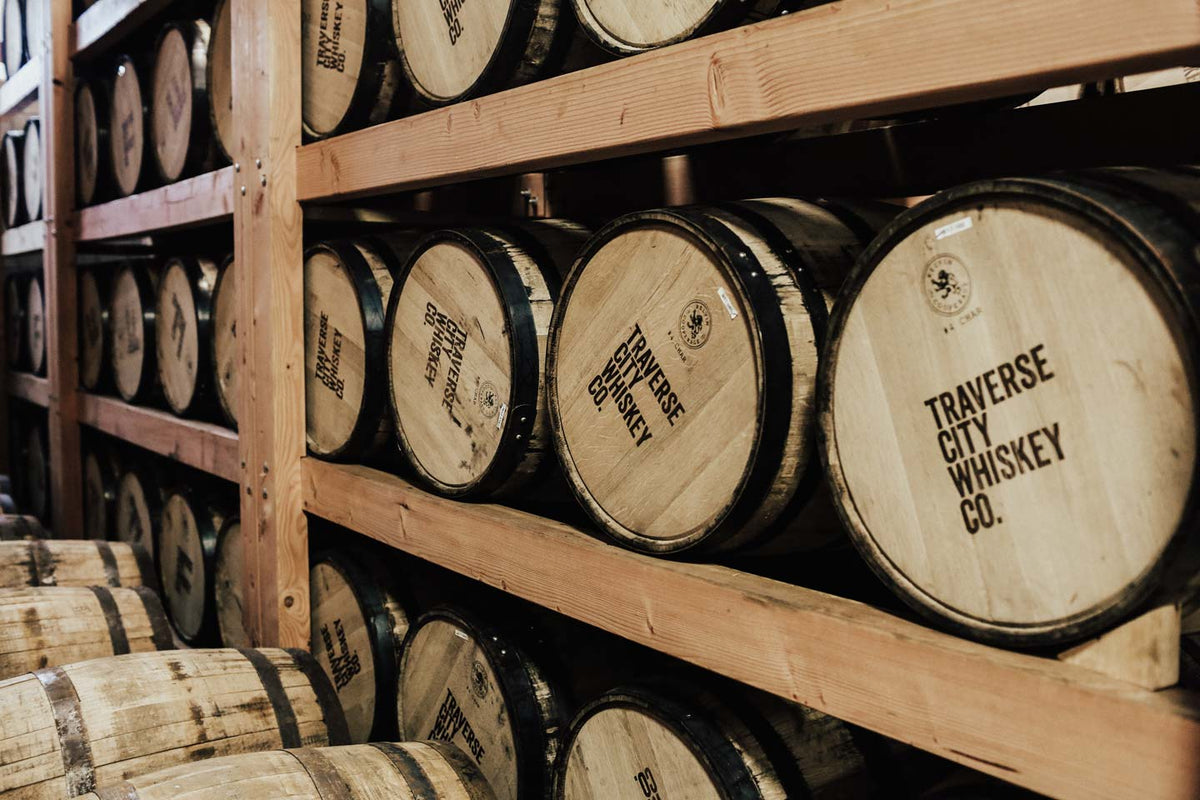 Wooden whiskey barrels stacked on wooden shelves in a warehouse, with one barrel clearly labeled TRAVERSE CITY WHISKEY CO.