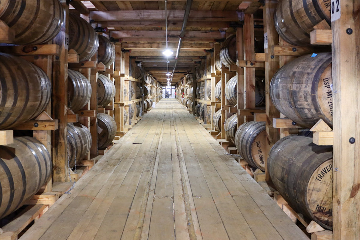 A long wooden walkway aisle between rows of large oak barrels on racks, likely inside a distillery or warehouse.