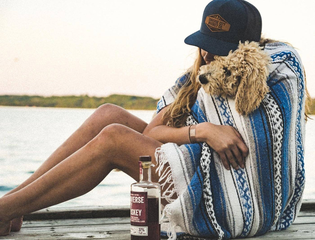 A woman sitting on a wooden dock by the water, wrapped in a blue and white patterned knit blanket and hugging a small fluffy dog. She is wearing a dark navy baseball cap with a brown logo patch.
