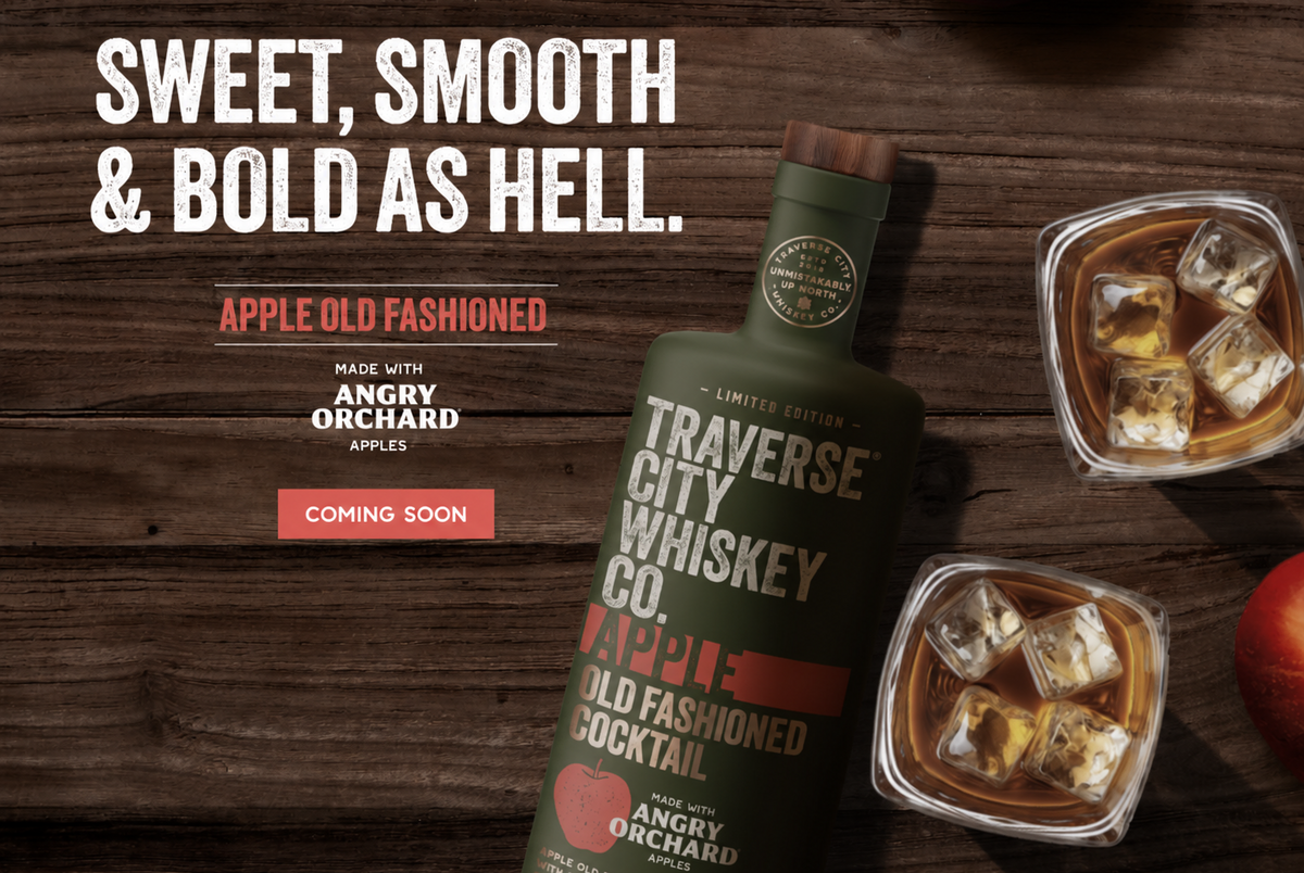 A dark green bottle of Traverse City Whiskey Co. Apple Old Fashioned Cocktail is displayed on a dark wood background next to two glasses filled with an amber cocktail and ice cubes, with promotional text on the left.