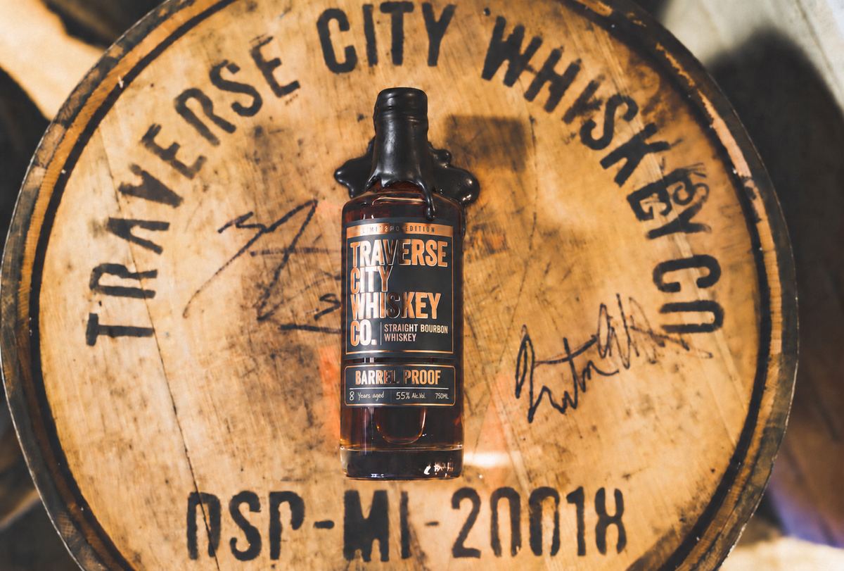 A bottle of Traverse City Whiskey Co. barrel-proof bourbon whiskey stands centered on a large wooden whiskey barrel with black printed text and signatures.