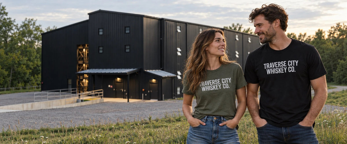 A man and a woman standing outdoors in front of a large black warehouse building, both wearing matching T-shirts with the text 'TRAVERSE CITY WHISKEY CO.' printed on the front.